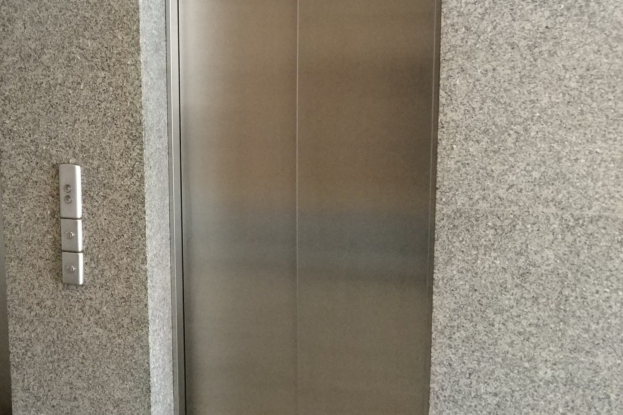 Geschlossene Aufzugstür aus gebürstetem Edelstahl in einer Granitwand, daneben die Ruftaster