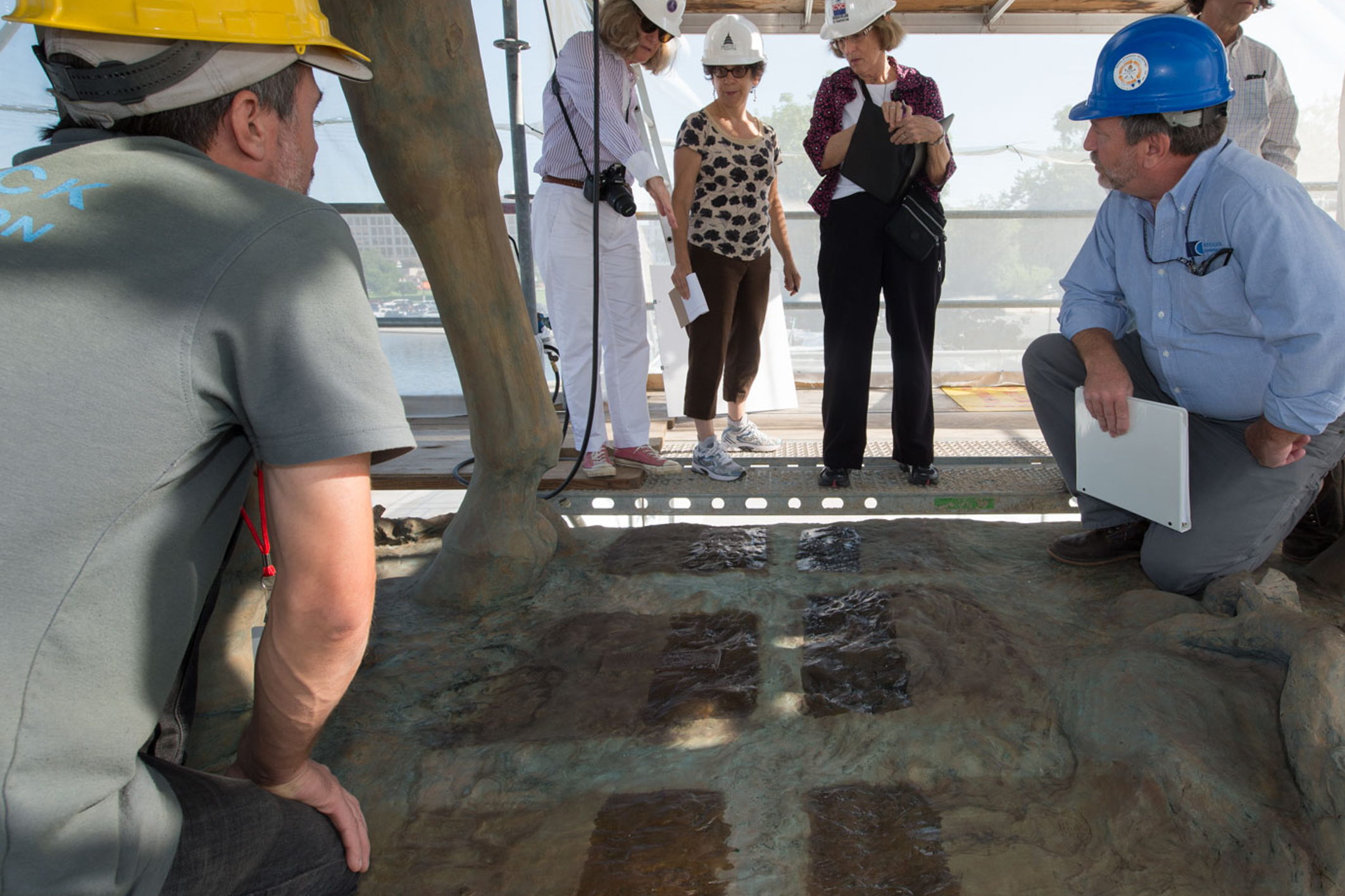 Fachleute mit Schutzhelmen begutachten auf einem Gerüst abgegrenzte Musterfelder auf dem Sockel einer Bronzeskulptur