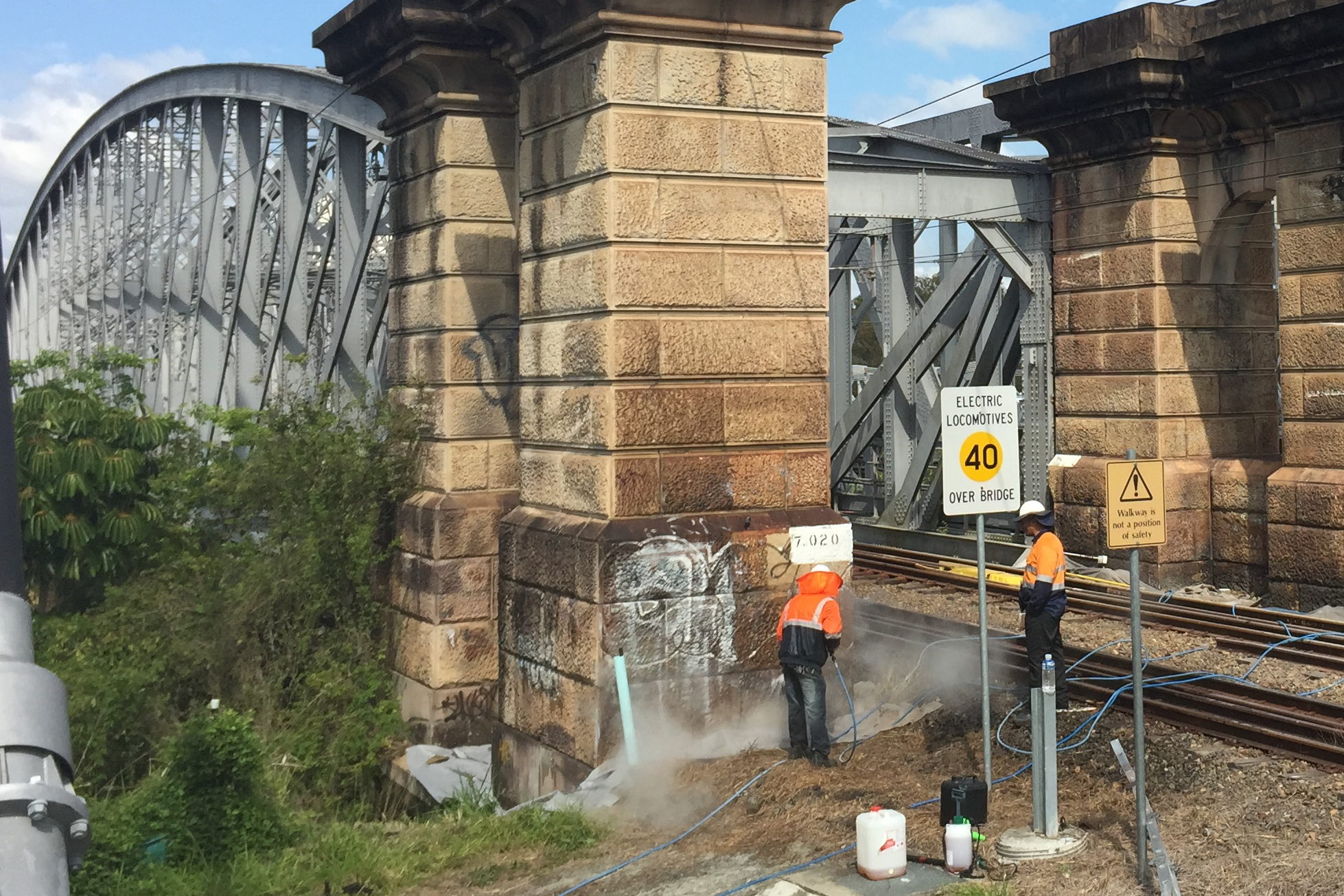 Zwei Arbeiter in Warnkleidung entfernen Graffiti von einem Sandsteinpfeiler einer Eisenbahnbrücke