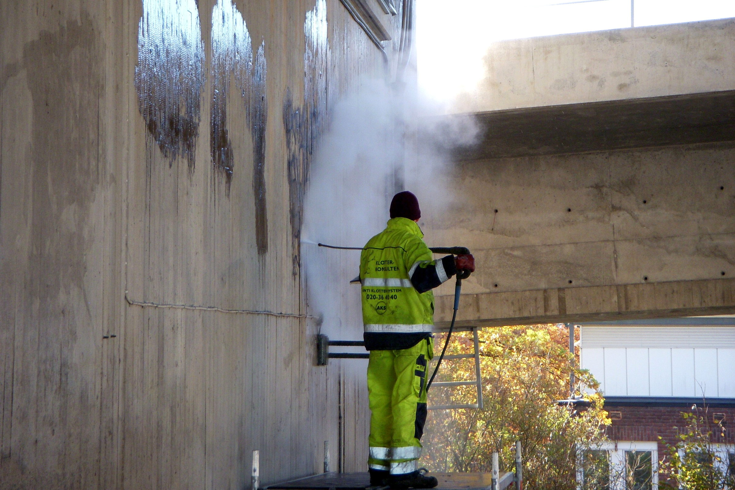 Arbeiter in Warnkleidung reinigt von einem Podest aus eine Betonwand mit Graffiti-Resten unter einer Brücke, Dampf steigt auf