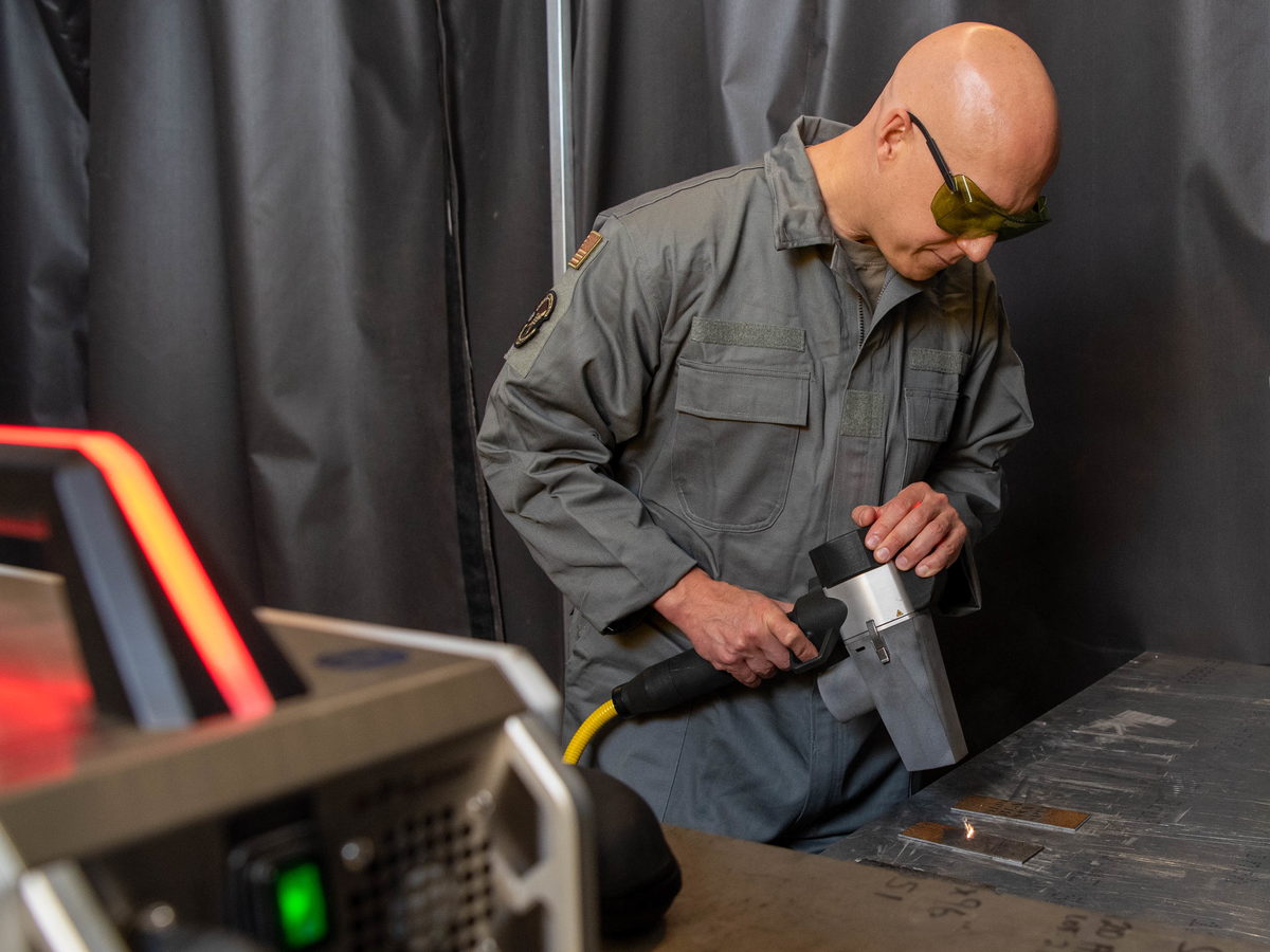 Techniker mit Schutzbrille führt einen Handlaser über eine lackierte Metallplatte, daneben die Lasereinheit