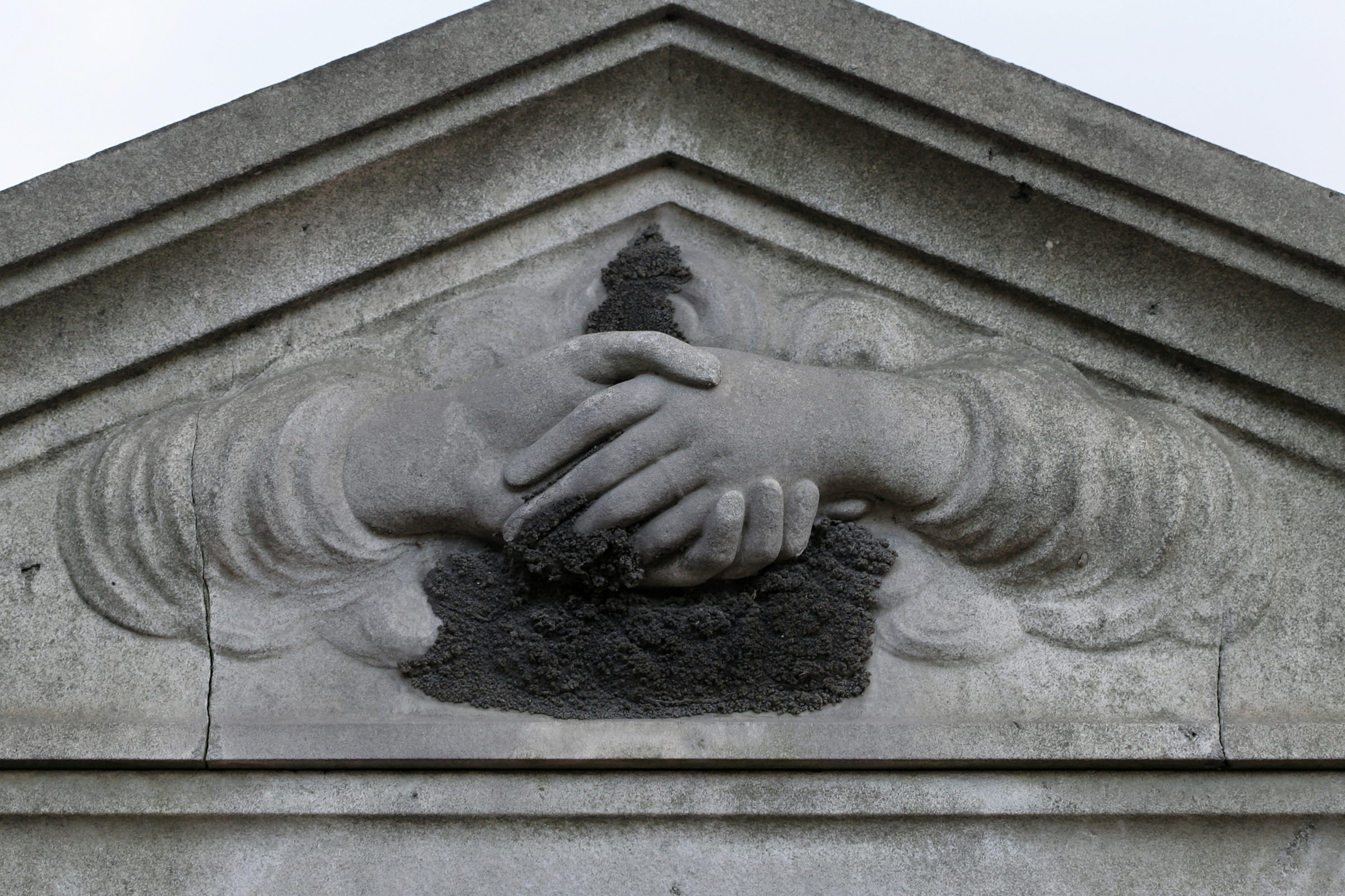 Steinrelief zweier verschränkter Hände in einem Giebel, in den Vertiefungen sitzt eine dunkle Kruste