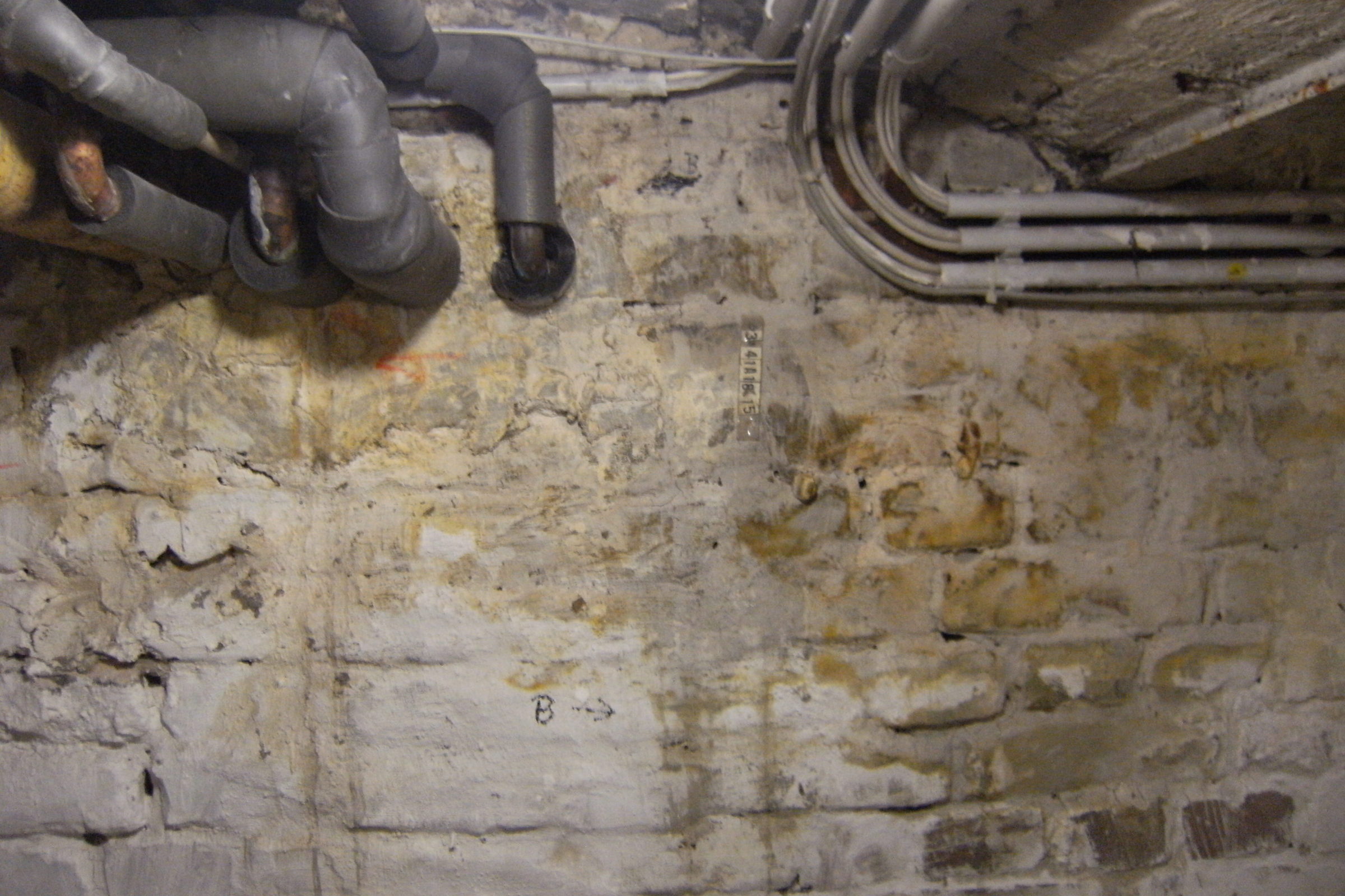 Kellerwand aus Mauerwerk mit Verfärbungen nach einem Wasserschaden, darüber Rohrleitungen und Kabel